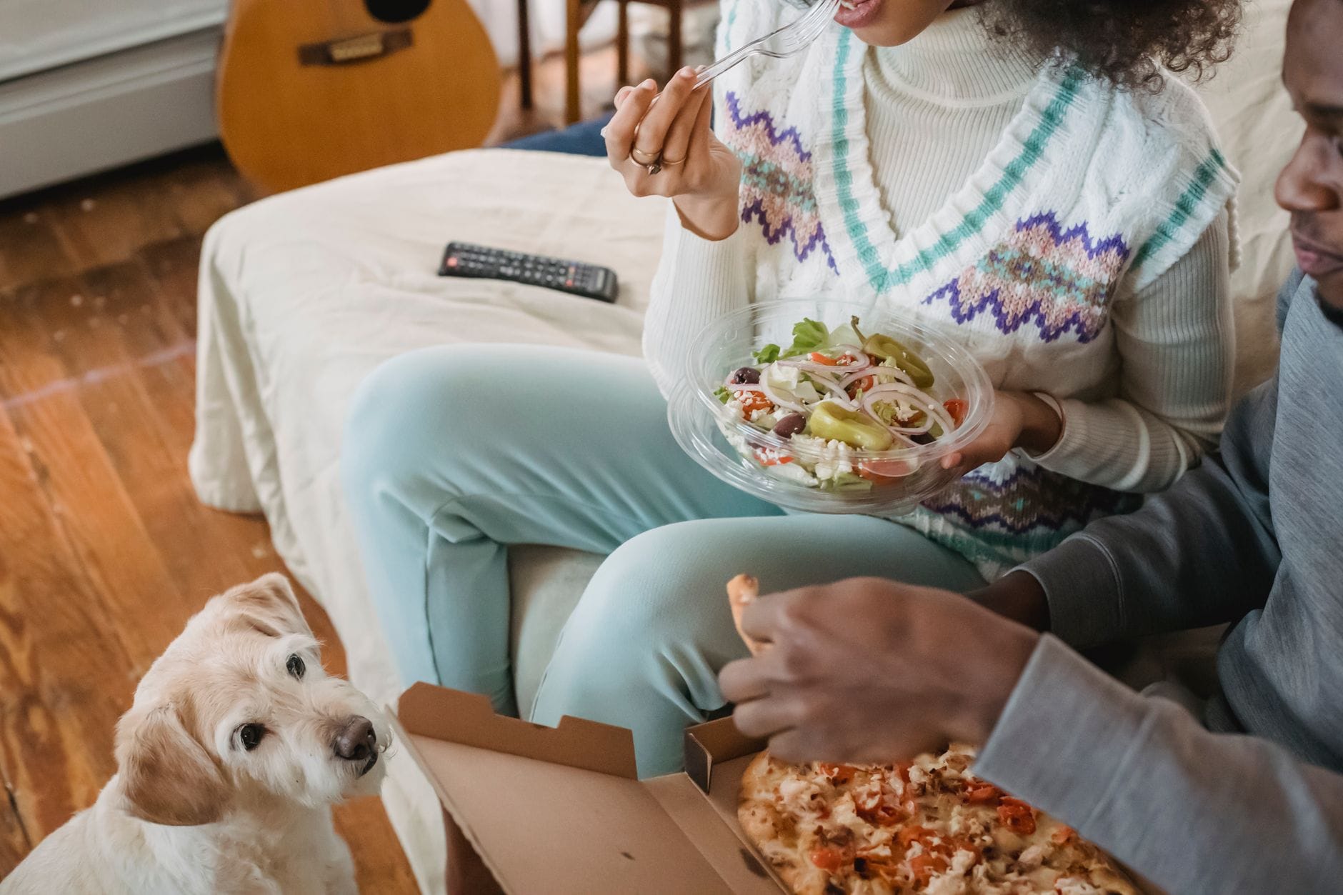 Para na kanapie je pizzę i sałatkę obok proszącego o jedzenie psa